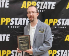 Chris Taylor holds the inaugural Hickory Innovation Award in front of a banner with repeating App State Hickory logo