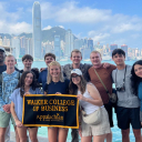 Holland Fellows from Appalachian State University hold a Walker College of Business banner along the Tsim Sha Tsui Promenade in front of the Victoria Harbour waterfront in Hong Kong. The International Finance Centre and the Hong Kong Island skyline are visible in the background. 