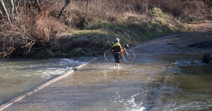 A cyclist crosses a flooded New River on foot, bike in hand.