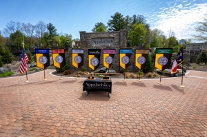 Displayed in front of the Appalachian State University sign, located in Founders Plaza on App State’s campus, are the university mace and banner, along with the banners for App State’s College of Arts and Sciences, Beaver College of Health Sciences, College of Fine and Applied Arts, Hayes School of Music, Reich College of Education, Walker College of Business and Cratis D. Williams School of Graduate Studies. The state flag is shown at far right and the U.S. flag is pictured at far left.