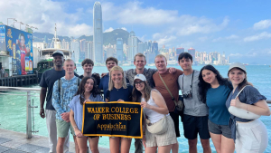 Holland Fellows from Appalachian State University hold a Walker College of Business banner along the Tsim Sha Tsui Promenade in front of the Victoria Harbour waterfront in Hong Kong. The International Finance Centre and the Hong Kong Island skyline are visible in the background. 