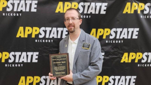 Chris Taylor holds the inaugural Hickory Innovation Award in front of a banner with repeating App State Hickory logo